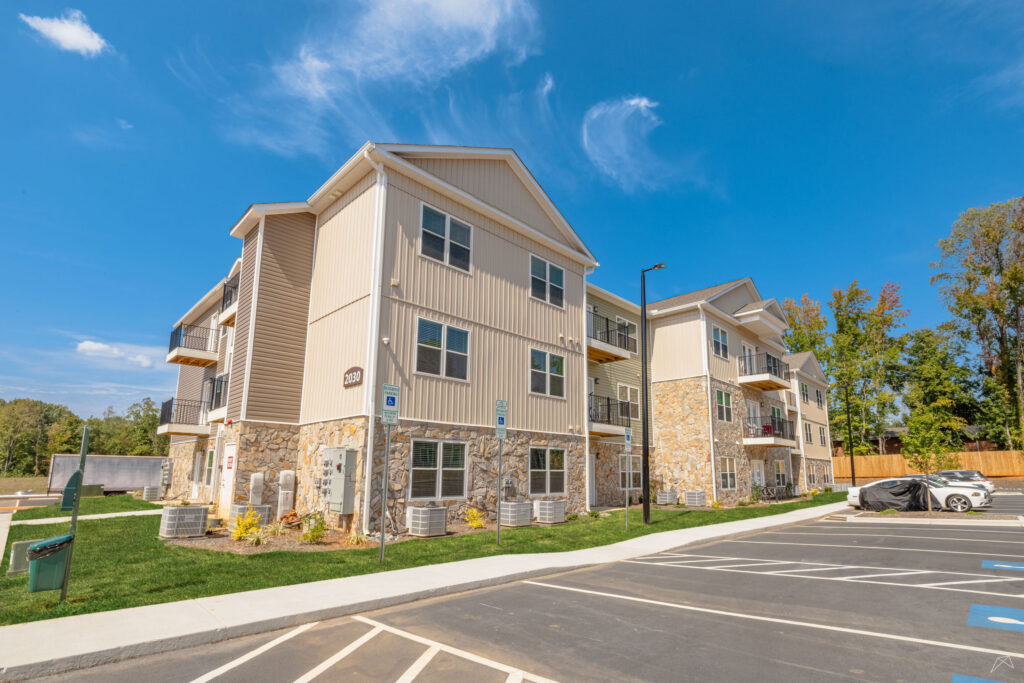 Exterior building of Preston Park Apartments in Burlington. Showing a green sage colored building with a parking lot and lush landscaping.