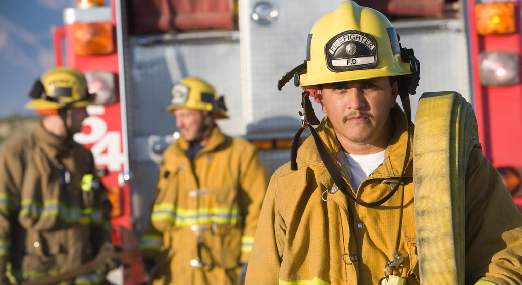 Firefighter in full protective gear and helmet standing in front of a fire truck, with fellow firefighters in the background.